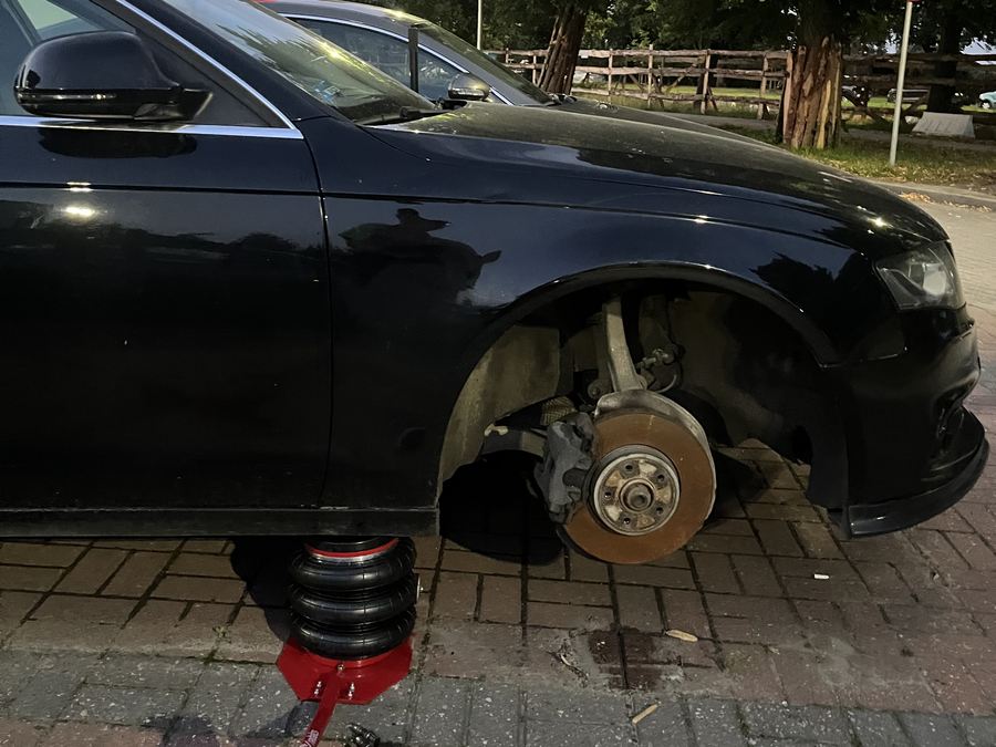 Czarne auto uniesione na pneumatycznym podnośniku przy krawężniku, zdjęte przednie koło i odsłonięta tarcza hamulcowa
