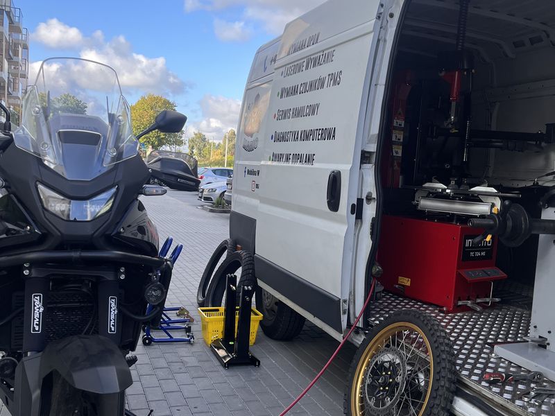 Otwarty bus serwisowy i motocykl na podnośniku, obok koło z felgą szprychową i rozłożone narzędzia